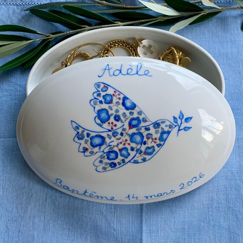 May include: White oval ceramic jewelry box with a blue floral dove design. The lid is inscribed with the name "Ad&egrave;le" and the date "14 mars 2026". The box is open, revealing gold jewelry inside.