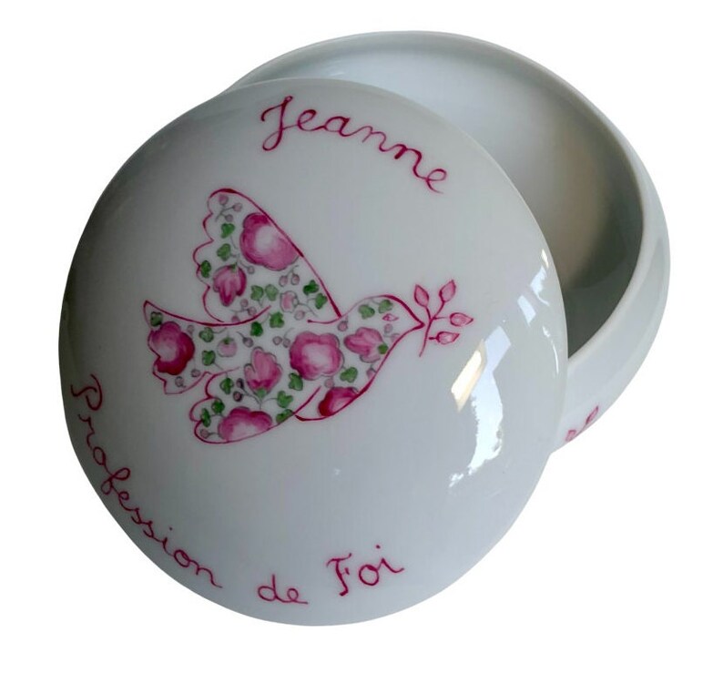 May include: A white porcelain trinket box with a lid. The lid features a dove design filled with pink flowers and green leaves, with the name "Jeanne" and the words "Profession de Foi" in red script.
