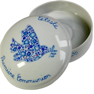 May include: White ceramic trinket box with a lid. The lid features a blue dove design with floral patterns and the words "Premi&egrave;re Communion" and "C&eacute;leste" in blue script. The box is open, revealing its interior.