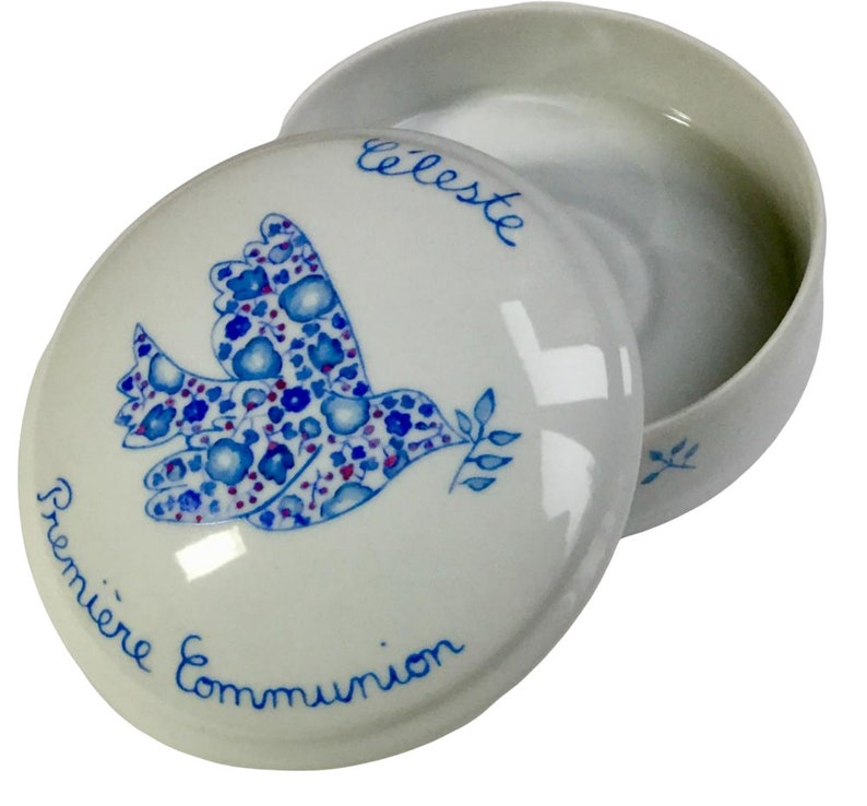 May include: White ceramic trinket box with a lid. The lid features a blue dove design with floral patterns and the words "Premi&egrave;re Communion" and "C&eacute;leste" in blue script. The box is open, revealing its interior.
