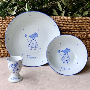 Peut inclure: Un ensemble de vaisselle pour enfants en céramique blanche avec une bordure bleue et un motif de souris fantaisiste. L'ensemble comprend une grande assiette, un bol plus petit et un gobelet à œufs. Chaque pièce présente une souris bleue tenant une étoile et le mot "Come" en lettres bleues.