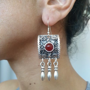 Könnte beinhalten: Silberfarbene Ohrringe mit einem quadratischen, verzierten Design. Jeder Ohrring hat einen zentralen, runden, roten Edelstein und drei hängende, tropfenförmige Anhänger. Die Ohrringe werden am Ohr einer Person getragen.