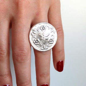 Könnte beinhalten: Ein silberner Ring mit einem großen, runden, verzierten Design. Der Ring wird am Mittelfinger einer Hand mit rotem Nagellack getragen.