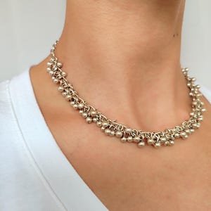 Op de afbeelding: Zilverkleurige ketting met kleine, ronde kralen die bij elkaar gegroepeerd zijn. De ketting wordt om de hals gedragen, tegen een witte top. De kralen zijn gelijkmatig verdeeld en creëren een delicate, getextureerde uitstraling.