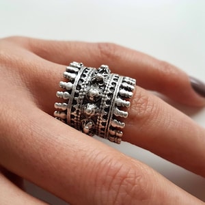 Könnte beinhalten: Ein silberfarbener Ring mit einem verzierten Design, das mehrere Reihen strukturierter Muster und kleine, erhabene Verzierungen aufweist. Der Ring ist breit und bedeckt einen großen Teil des Fingers, mit einem detaillierten, Vintage-Look.