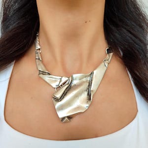 May include: A silver-toned statement necklace with a unique, abstract design. The necklace features a large, textured central pendant with folded and layered metal elements. The chain is delicate, and the overall style is modern and artistic.