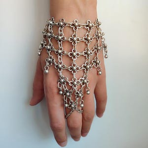 Könnte beinhalten: Silberfarbenes Handkettenarmband. Das filigrane Design weist ein netzartiges Muster mit floralen Akzenten und hängenden Perlen auf. Das Armband enthält einen Ring, der mit der Handkette verbunden ist, was ein einzigartiges Accessoire ergibt.
