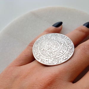 May include: A silver ring with a large, round, ornate design. The ring is worn on a finger.