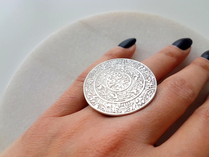 May include: A silver ring with a large, round, ornate design. The ring is worn on a finger.