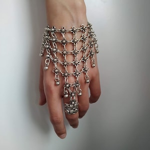 Könnte beinhalten: Ein silbernes Handkettenarmband mit floralem Design. Das Armband hat ein netzartiges Muster mit hängenden Perlen und einem Ring, der mit der Kette verbunden ist. Der Schmuck wird an einer Hand vor weißem Hintergrund getragen.