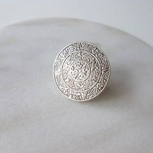 May include: A silver ring with a circular design featuring intricate details and a central floral motif.
