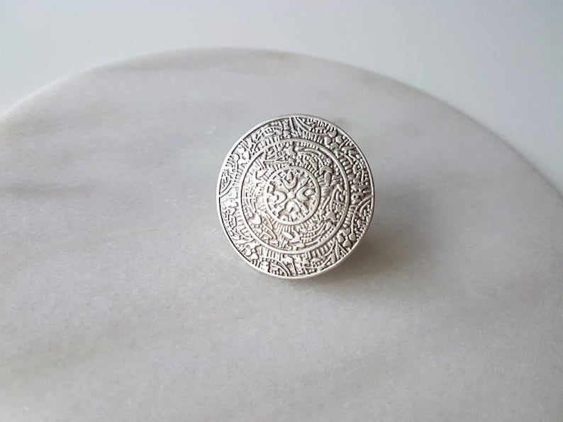 May include: A silver ring with a circular design featuring intricate details and a central floral motif.