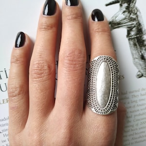Könnte beinhalten: Ein silberner Ring mit ovalem Mittelteil und detailliertem Rand wird an einem Finger mit schwarzem Nagellack getragen. Der Ring hat einen antiken oder Vintage-Stil.