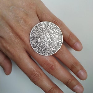 May include: A large silver ring with an intricate, circular design. The ring is worn on a person's finger.