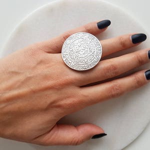 May include: A large silver ring with an intricate design. The ring is worn on a finger with black nail polish.