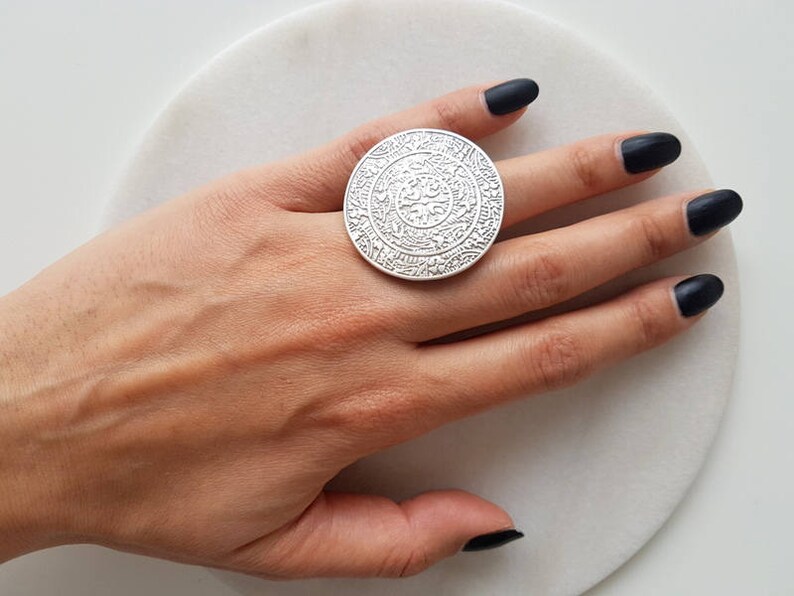 May include: A large silver ring with an intricate design. The ring is worn on a finger with black nail polish.