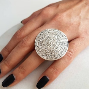 Könnte beinhalten: Ein großer silberner Ring mit einem komplizierten, kreisförmigen Design. Der Ring wird an einem Finger mit schwarzem Nagellack getragen.