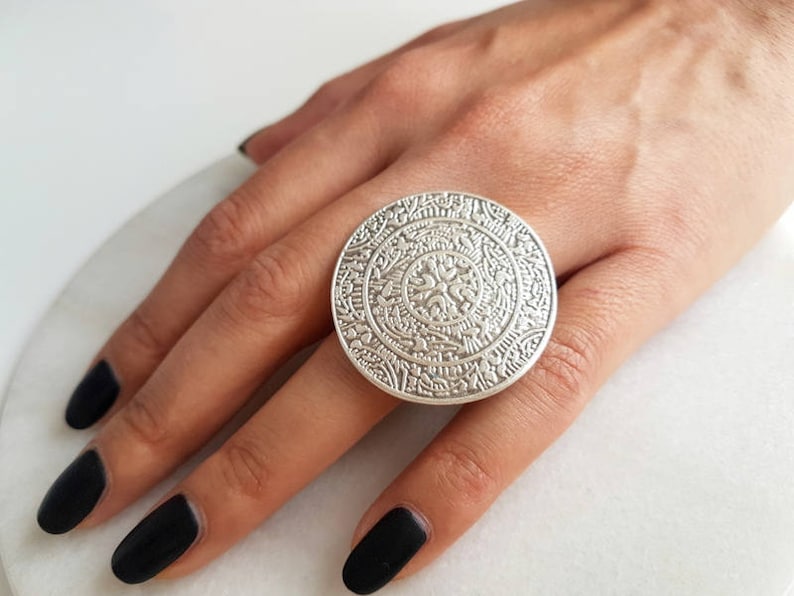 May include: A large silver ring with an intricate, circular design. The ring is worn on a finger with black nail polish.