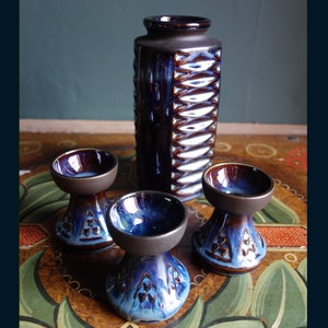 Puede incluir: Conjunto de cerámica en tonos azules y marrones. La imagen muestra un jarrón alto y rectangular con un diseño texturizado, junto con tres cuencos pequeños con pedestal. Los artículos tienen un acabado brillante.