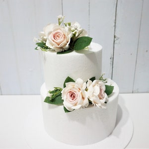 May include: Two-tiered white cake decorated with pink roses and greenery. The cake is on a white cake stand.