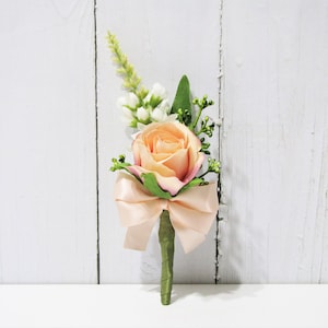 May include: Peach-colored rose with white flowers and greenery, tied with a peach-colored satin ribbon. This boutonniere is perfect for a wedding or special occasion.