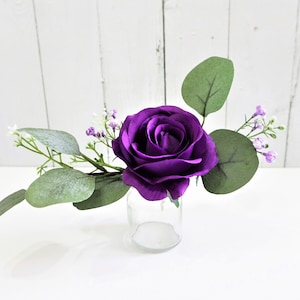 May include: A single purple rose with small white and purple flowers and green leaves in a clear glass vase.