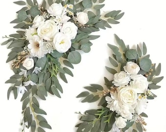 May include: Two white floral arrangements with greenery. The flowers are white roses, hydrangeas, and other small white flowers. The greenery is eucalyptus leaves.