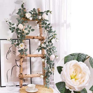 Op de afbeelding: Een houten ladder is versierd met witte rozen slingers en groene bladeren. De rozen staan in volle bloei, met een close-up van een enkele bloem. Een ingelijste kunstafdruk en een theekopje zijn zichtbaar op de achtergrond.