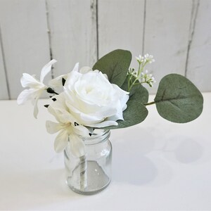 May include: A small white rose, white flowers, and green eucalyptus leaves arranged in a clear glass jar.