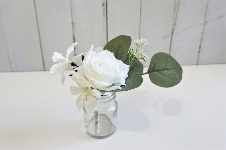May include: A small white rose, white flowers, and green eucalyptus leaves arranged in a clear glass jar.
