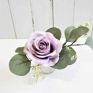 May include: A single, light purple silk rose with green eucalyptus leaves and small white flowers in a clear glass vase.