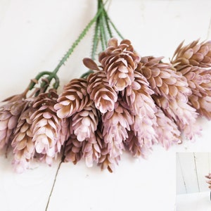 Puede incluir: Un ramo de flores artificiales de color marrón claro y rosa-púrpura con tallos verdes. Las flores tienen forma de cono y están dispuestas en un ramo. El fondo es una superficie de madera blanca. Un pequeño jarrón negro con flores está en la esquina inferior derecha.