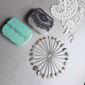 May include: A set of 20 silver and gold decorative pins with various floral designs. The pins are arranged in a circle on a gray surface. Two small silver boxes with lace designs are also in the image.