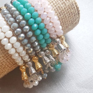 Op de afbeelding: Een stapel kleurrijke kralenarmbanden met gouden en zilveren kwastcharms. De armbanden zijn gemaakt van turquoise, grijze, witte en roze kralen.