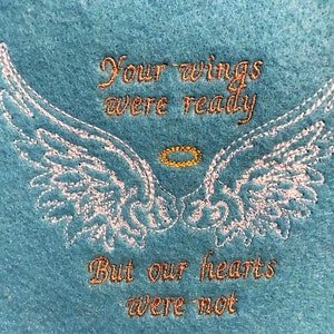 Könnte beinhalten: Ein blaugrüner Stoff mit weißen gestickten Engelsflügeln und einem goldenen Heiligenschein. Der Text "Deine Flügel waren bereit, aber unsere Herzen waren es nicht" ist in Gold gestickt.