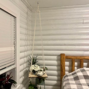 May include: A white macrame plant hanger with a wooden shelf and three potted plants. The shelf is hanging from a hook on the ceiling. The shelf is decorated with a white toy truck. The shelf is in a room with white walls and a wooden bed with a gray and white checkered blanket.