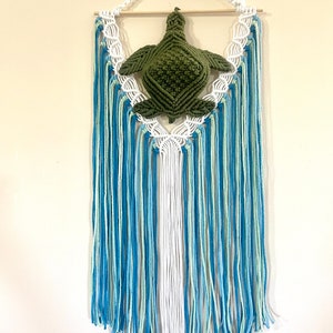 May include: A green macrame turtle hanging from a wooden dowel with blue, white, and green yarn tassels.