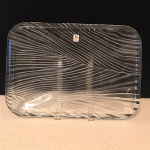 May include: Clear rectangular glass tray with a wave-like pattern. The tray has rounded corners and a small label in the upper right corner. The tray is displayed on a white surface with a black background.