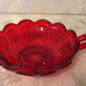 May include: A red glass bowl with a scalloped edge and a handle. The bowl has a textured design with small circles.