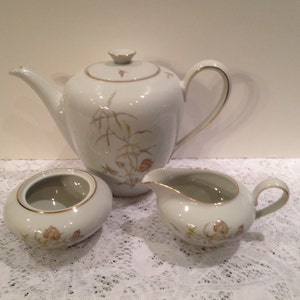 May include: A white porcelain tea set with a teapot, sugar bowl, and creamer. The set features gold trim and a delicate floral and wheat design in shades of gold and brown. The teapot has a curved spout and handle.