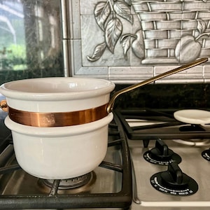 May include: A white ceramic double boiler pot featuring a polished copper band around the center and a long, slender metallic handle extending from the side. The pot has a smooth, rounded body and a wide rim, designed for heating contents.
