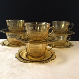 May include: Set of six amber glass teacups and saucers with a floral design. The cups and saucers are stacked on top of each other.