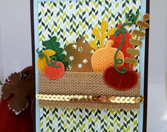 May include: A handmade autumn-themed greeting card featuring paper pumpkins in shades of orange, yellow, and brown. The pumpkins are arranged on a burlap strip with gold sequins, set against a patterned background of green, blue, and white chevrons.