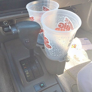May include: Two clear plastic cups with lids are in a car cup holder. The cups have the words "KUDER BIG Gulp" printed on them. The car's gear shift is visible in the background.