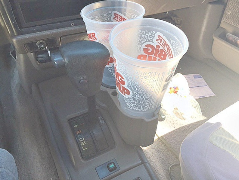 May include: Two clear plastic cups with lids are in a car cup holder. The cups have the words "KUDER BIG Gulp" printed on them. The car's gear shift is visible in the background.