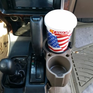 May include: A close-up of a car's interior with a cup holder and a cup of soda. The cup holder is made of plastic and is grey in colour. The cup is white with a red, white and blue American flag design and the text "Carl's Jr." printed on it.