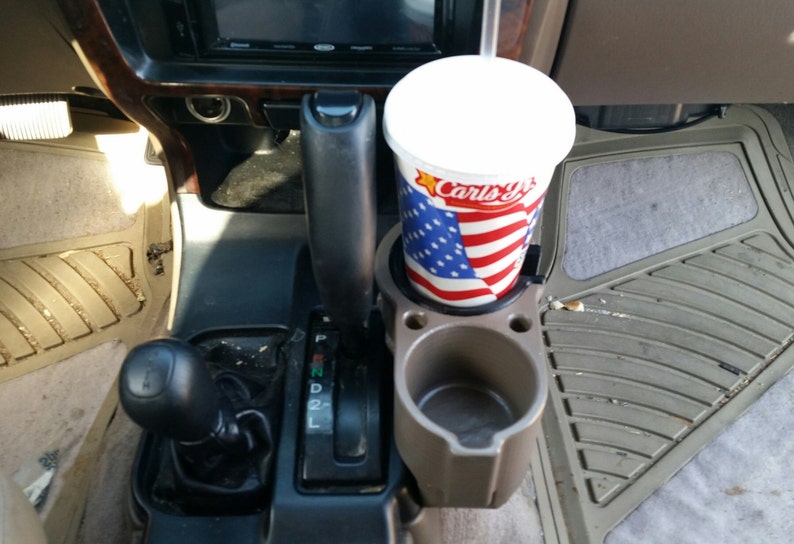 May include: A close-up of a car's interior with a cup holder and a cup of soda. The cup holder is made of plastic and is grey in colour. The cup is white with a red, white and blue American flag design and the text "Carl's Jr." printed on it.
