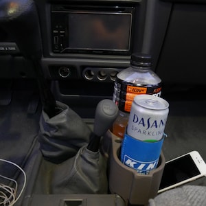 May include: A close-up of a car's interior with a cup holder containing a can of Dasani Sparkling water and a clear plastic bottle of ION+ sports drink. A cell phone is in the cup holder next to the can.