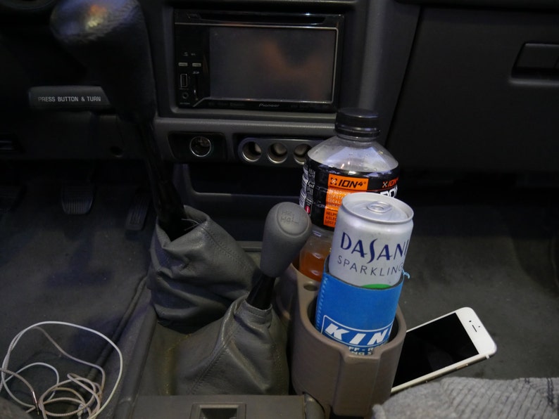 May include: A close-up of a car's interior with a cup holder containing a can of Dasani Sparkling water and a clear plastic bottle of ION+ sports drink. A cell phone is in the cup holder next to the can.