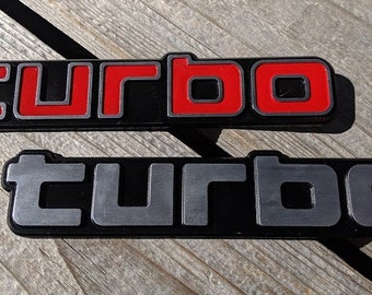 Może przedstawiać: Dwa czarne i srebrne emblematy "turbo". Górny emblemat ma czerwone litery.
